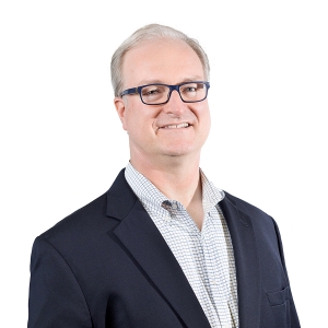 A smiling individual wearing glasses, a dark blazer, and a patterned shirt against a solid white background.