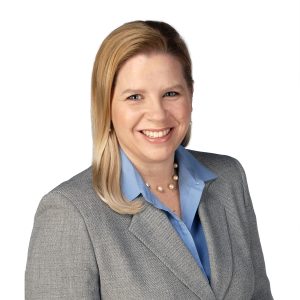 A smiling person wearing a gray blazer, light blue shirt, and a pearl necklace.