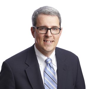 A smiling person wearing glasses, a dark blazer, a light blue striped tie, and a white shirt against a light background.