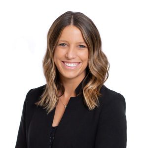 A smiling person with wavy hair wearing a black blazer stands against a white background.
