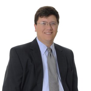 A person is wearing a dark suit jacket, a light-colored shirt, and a dotted tie, and is smiling against a plain background.