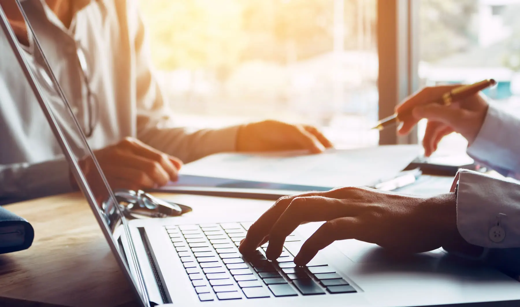 Individuals are working on a laptop at a desk with a sunlit background, indicating a business or educational setting.