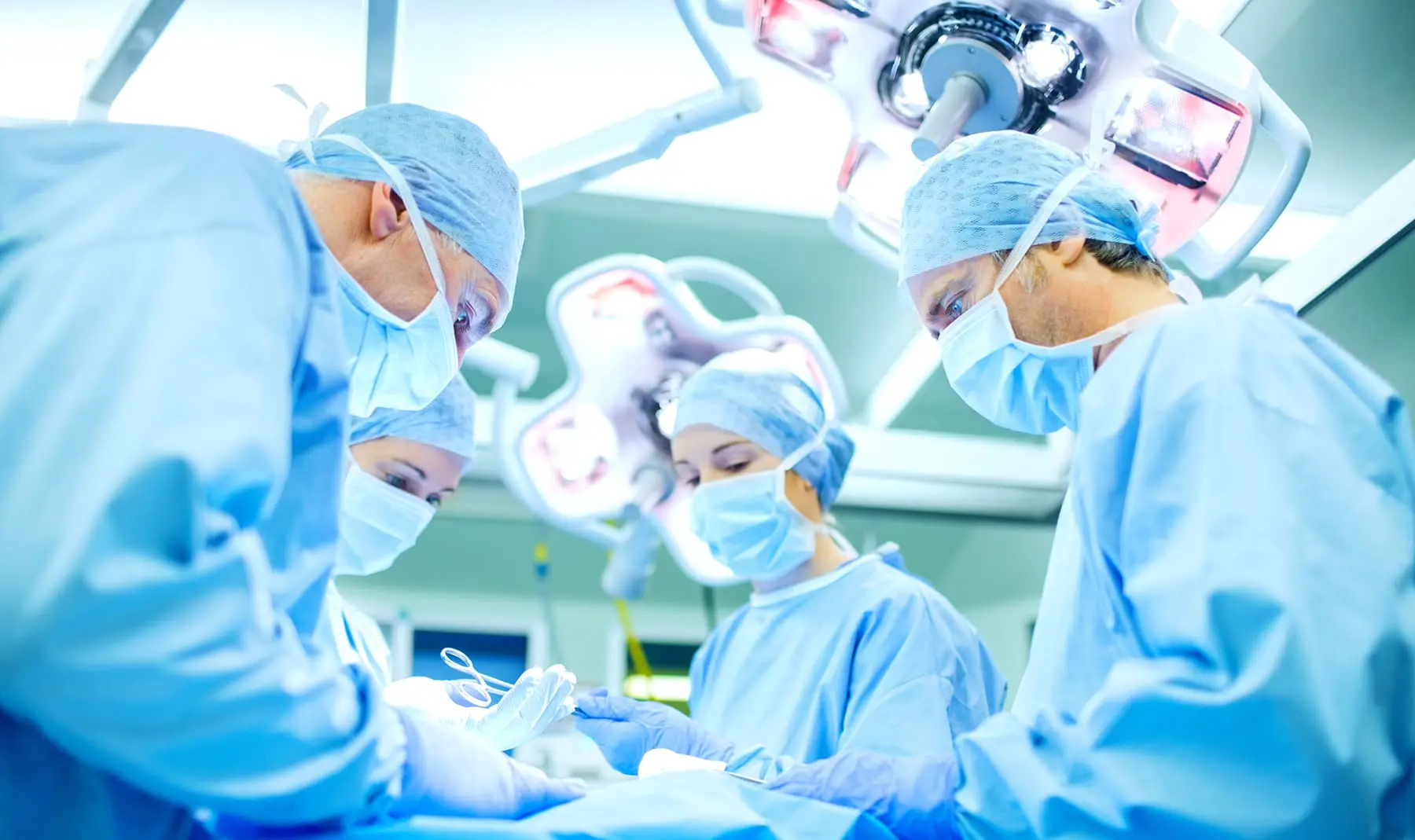 Medical personnel in scrubs and surgical masks are focused on a procedure under bright operating room lights.