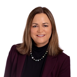 A smiling woman with shoulder-length hair is wearing a black turtleneck, a maroon blazer, and a pearl necklace.
