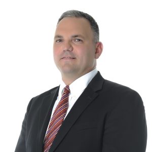 A person with short hair is wearing a dark suit jacket with a white shirt and a striped tie, posing in front of a white background.