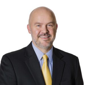 A man with a bald head, beard, and mustache is smiling while wearing a suit with a yellow tie against a white background.