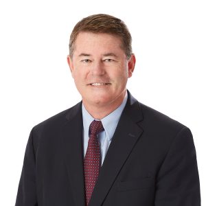 A person is wearing a dark suit, white shirt, and a dotted tie, with a smile against a white background.