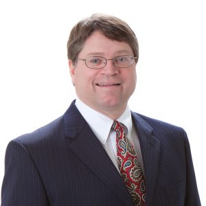 A smiling person wearing glasses, a navy pinstripe suit, a white shirt, and a paisley tie with a white background.
