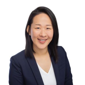 A person with dark hair is wearing a navy blazer and white shirt while smiling against a white background.
