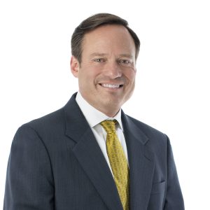 A smiling individual wearing a dark suit, white shirt, and yellow tie stands against a white background.