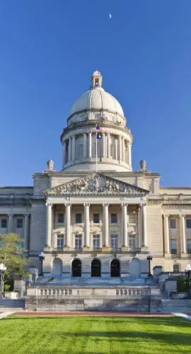 A grand neoclassical building with a large dome is basked in sunlight against a clear blue sky, surrounded by immaculate lawns and walking paths.