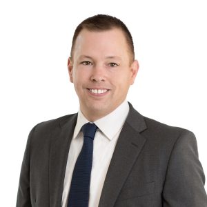 A person is wearing a gray suit with a blue tie and smiling at the camera against a white background.