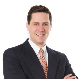 A smiling person dressed in business attire with a striped orange tie and crossed arms against a white background.