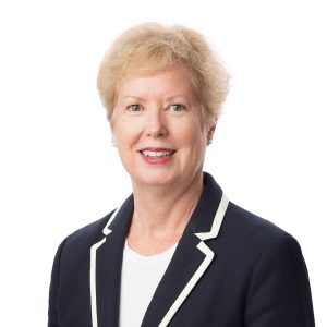 A smiling person with short blonde hair wearing a dark jacket with white piping and a light-colored shirt against a white background.