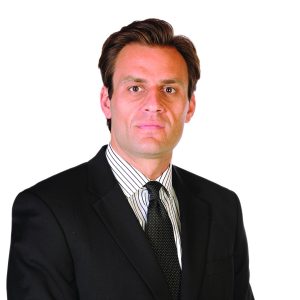 A person wearing a black suit and striped tie stands against a white background.