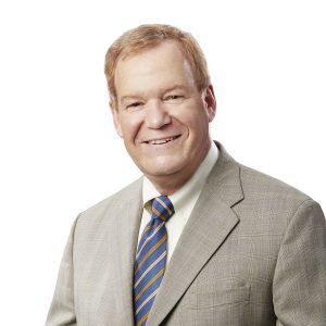 A person with short hair is smiling and wearing a light gray suit with a striped blue and yellow tie against a white background.