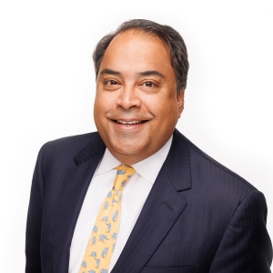 A person wearing a dark suit, white shirt, and yellow tie smiles against a white background.