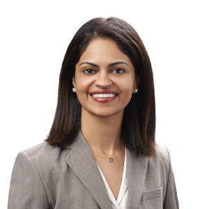 A person with shoulder-length dark hair wearing a grey blazer and a white blouse is smiling against a white background.
