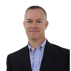 A smiling person dressed in a business suit with a checkered shirt stands against a plain white background.
