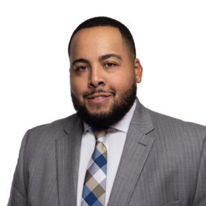 A man with a beard is wearing a light gray suit, white shirt, and plaid tie, smiling with a white background.