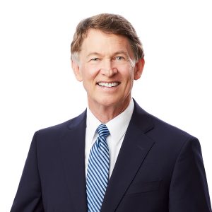 A smiling person dressed in a professional navy blue suit with a light blue striped tie and a white shirt against a white background.