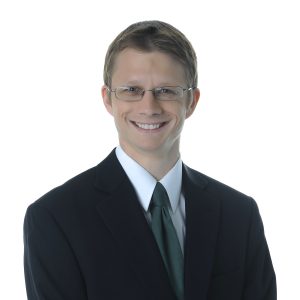A smiling individual wearing glasses, a black suit, white shirt, and a green tie against a white background.