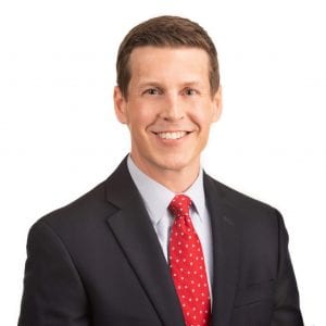 A man with short brown hair is smiling and wearing a dark suit with a light blue shirt and red tie.