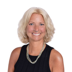 A woman with blonde hair is smiling while wearing a black sleeveless top and layered necklaces.