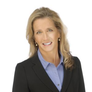 A smiling person with blond hair wearing a black blazer and a blue shirt against a white background.
