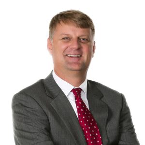 A smiling person wearing a gray suit and a red dotted tie stands against a white background.