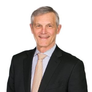 A man in a black suit, light-colored dress shirt, and beige tie smiles against a white background.