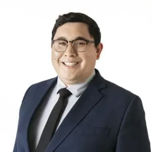 A person wearing glasses and a blue suit with a black tie smiles against a plain white background.