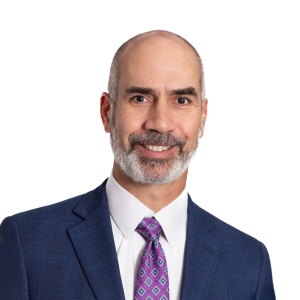 A person with a slight smile wearing a blue suit jacket and a purple patterned tie against a white background.