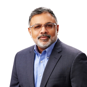 A person with graying hair and a beard is wearing glasses, a blue shirt, and a dark blazer, posing against a white background.