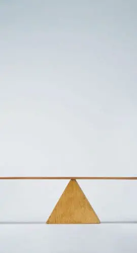 A piggy bank is balanced on a seesaw against a jar filled with coins and accompanied by small objects.