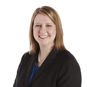 A smiling woman dressed in professional attire with a black blazer and a blue top, adorned with a pearl necklace, against a plain background.