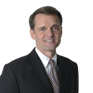 A smiling person wearing a suit with a tie stands against a plain background.