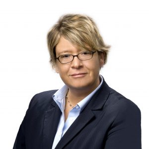 A person with short hair is wearing glasses, a dark blazer, and a light blue shirt.