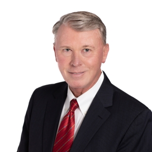 A person wearing a dark suit jacket, a white shirt, and a red tie poses for a professional portrait with a white background.