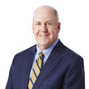 A person wearing a dark suit, white shirt, and a yellow and blue striped tie with a white background.