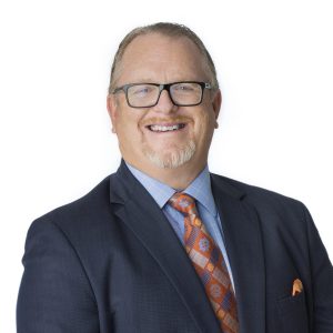 A smiling person wearing glasses is dressed in professional attire with a tie and suit jacket.