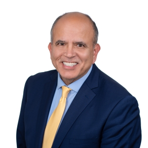 A person wearing a dark blue suit, light blue shirt, and yellow tie is smiling against a white background.