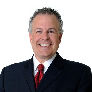 A smiling individual with curly hair dressed in a business suit with a red tie against a white background.