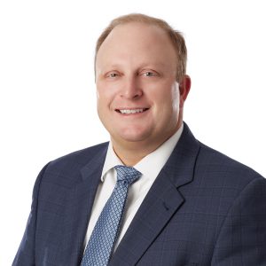 A smiling individual wearing a business suit and tie with a white shirt on a neutral background.