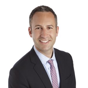A person is smiling for a professional headshot, wearing a black suit, a white shirt, and a pink patterned tie.