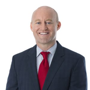 A smiling individual is dressed in professional attire with a red tie and a blue suit against a white background.