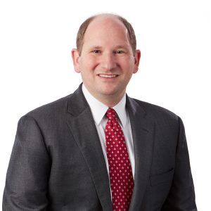 A smiling individual wearing a gray suit and a red tie with a white background.