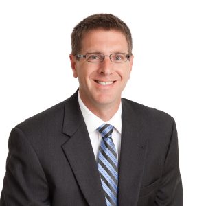 A smiling individual wearing glasses, a dark suit, a light blue striped tie, and a white shirt poses for a professional portrait against a plain background.
