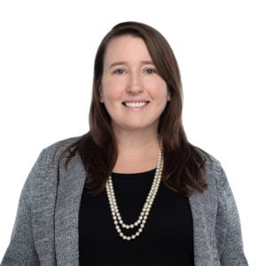 A person with long brown hair is smiling while wearing a black top, a grey cardigan, and a pearl necklace against a white background.