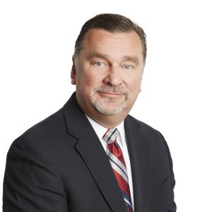 A person wearing a dark suit and a patterned red tie with a neatly trimmed beard is posing against a white background.